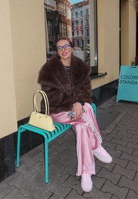 Malli ruskeassa tekoturkistakissa, pinkeissä satiinihousuissa, joissa valkoiset raidat, pinkit lenkkarivaljaat ja keltainen käsilaukku kädessä. Istuu turkoosilla penkillä.