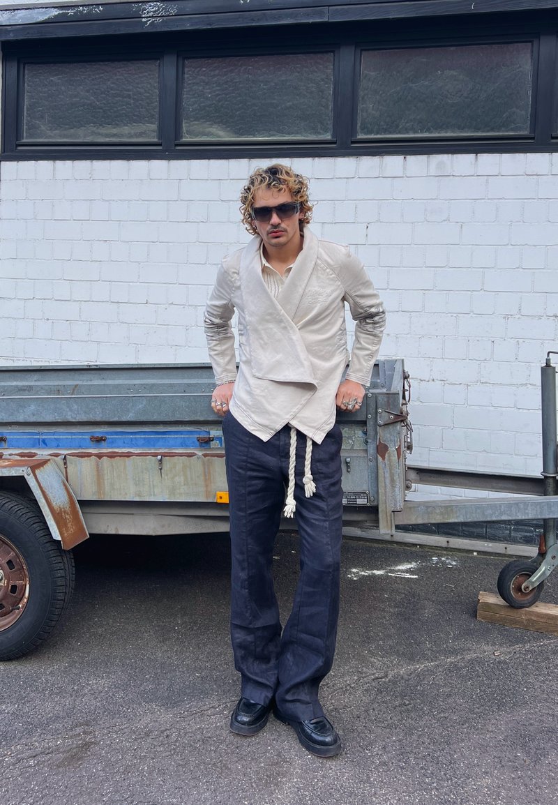 Homme aux cheveux bouclés et lunettes de soleil, debout devant une remorque rouillée, portant une veste cache-cœur beige avec ceinture en corde et un pantalon large sombre.