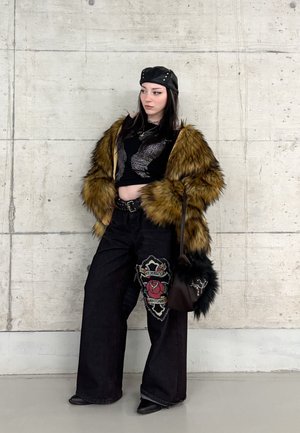 Joven con chaqueta de piel sintética, top corto negro, pantalones de pierna ancha con parche de rosa, sombrero de cuero y bolso negro de pelo.