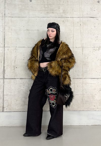 Joven con chaqueta de piel sintética, top corto negro, pantalones de pierna ancha con parche de rosa, sombrero de cuero y bolso negro de pelo.