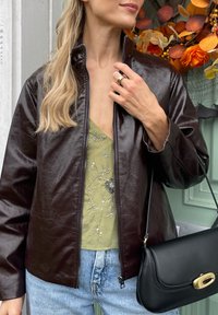 Brown faux leather jacket with a high collar, worn over a green top featuring floral embellishments, paired with light blue jeans and a black handbag.