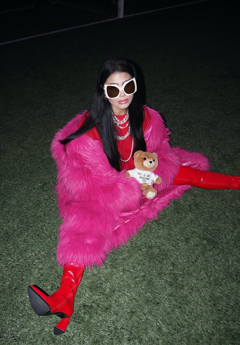 A woman sitting on the grass, sporting large white sunglasses, a pink fur coat, and red boots, while holding a teddy bear dressed in a white shirt.