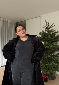 Sort plyssede frakke over en glitrende sort heldragt. Heldragten har en tætsiddende form og subtil glans. Grøn gaveæske med rødt bånd i baggrunden.