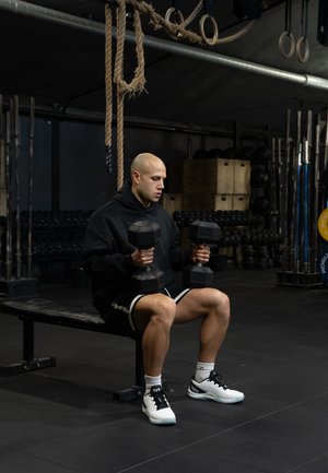 Black dumbbells held by a person in a black hoodie and shorts, sitting on a bench in a gym with dark flooring and a strength training area.