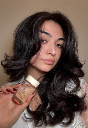Mujer con cabello largo y ondulado de color oscuro sostiene una pequeña botella del producto capilar Kérastase Gloss Absolu Bain Hydra-Glacé sobre un fondo neutro.