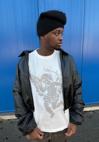 Fekete, fényes bomberdzseki fehér póló felett, rajta szürke angyalszárny grafika. Sötét hajú férfi fekete kötött sapkában.
