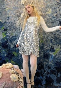 Robe en sequins argentés à coupe ample, associée à des collants blancs texturés et des talons hauts noirs, sur un fond texturé bleu.