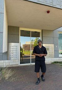 Camicia nera a maniche corte con bottoni e pantaloni corti, clogs neri, in piedi davanti a una porta di vetro con un pannello metallico per la posta.