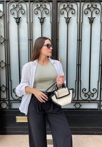 Vrouw met lang bruin haar die zwarte zonnebril draagt, een wit shirt over een grijze tanktop en zwarte broek, en een kleine beige en zwarte handtas vasthoudt.