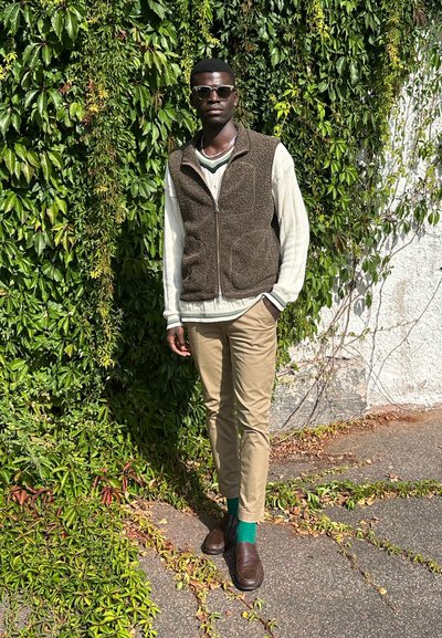 Chaleco de felpa marrón sobre una camiseta de manga larga crema, pantalones beige, calcetines verdes y mocasines oscuros, de pie contra un fondo de hiedra verde.
