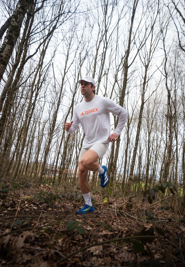 Δρομέας με λευκό μπλουζάκι Terrex, λευκό καπέλο και μπλε παπούτσια τρέχει σε μονοπάτι δάσους με γυμνά δέντρα και πεσμένα φύλλα.