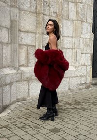 Vinrød pelsfrakke af imiteret pels med et voluminøst design, kombineret med en sort lang kjole og sort ankelhøje støvler med hæl på brosten.