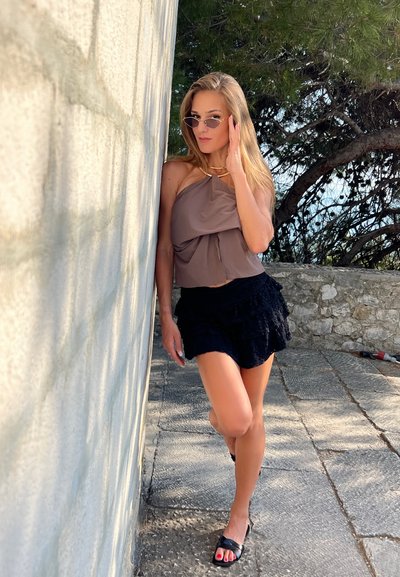 Top de halter marrón, falda negra con volantes y sandalias negras. La modelo está de pie contra una pared de piedra texturizada, con gafas de sol y cabello largo.