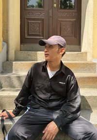 Svart skjorta med knappar och en vit logotyp, ihopparat med svarta träningsbyxor med ljusblå accenter. Sitter på stensteg.