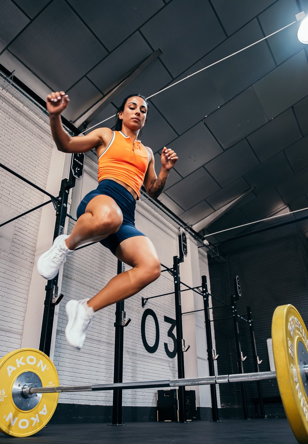 Sportists oranžā bezpieksnes tops un tumši zilo šortu komplektā, kas lecot pāri stieņiem ar dzeltenajiem svariem sporta zālē. Betona sienas un fitnesa aprīkojums redzami fona.