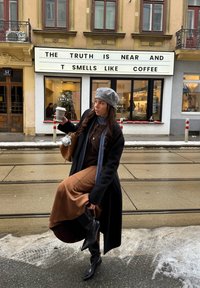 Femme vêtue d'habits d'hiver tenant une tasse de café, debout dans une rue enneigée devant un café avec une enseigne indiquant "La vérité est proche et elle sent le café."
