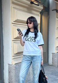 Λευκό T-shirt με μαύρες λεπτομέρειες και εκτύπωση "ΕΙΜΑΙ GCDS", συνδυασμένο με ανοιχτό μπλε τζιν. Τα αξεσουάρ περιλαμβάνουν σκούρα γυαλιά ηλίου και μαύρη τσάντα.