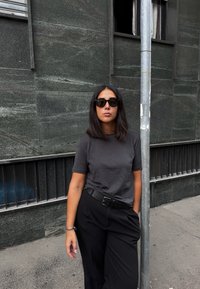 Ung kvinne med rett, mørkt hår og solbriller står med den ene hånden i lommen mot en mørk steinvegg nær en metallstang.