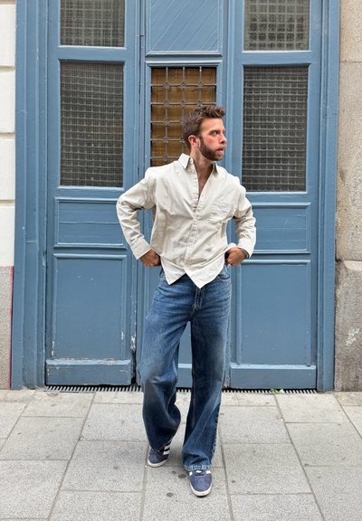 Hombre con camisa beige y vaqueros azules de pie en la acera, posando frente a grandes puertas dobles azules con paneles de vidrio.