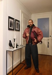 Vīna sarkana bomber jaka pār melnu kapuci, tumši platām biksēm, turot vīnogu krāsas mugursomu. Melnputns un sveces uz konsoles kā dekorējoši elementi.