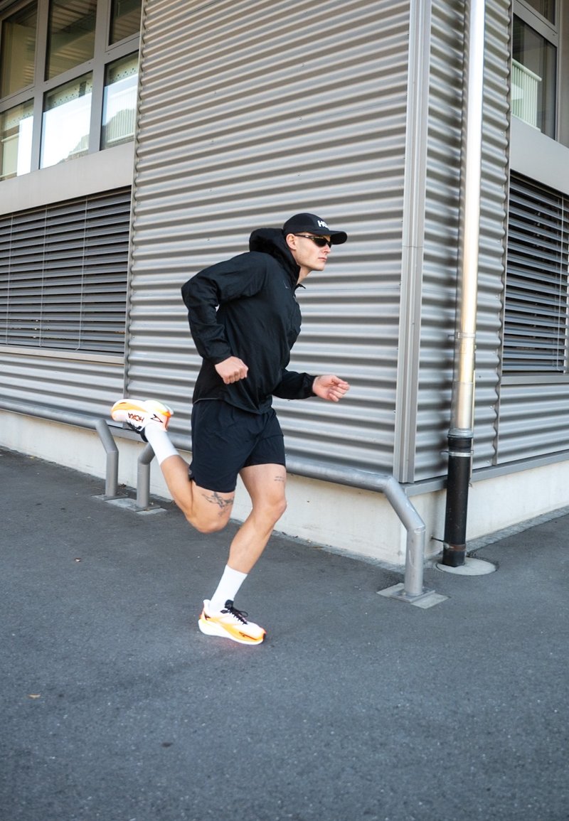 Black lightweight jacket, black shorts, and orange running shoes with black accents. Runner in motion near a corrugated wall.