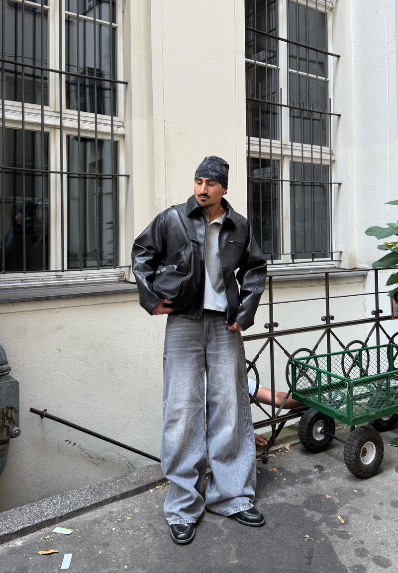 Homme en blouson en cuir noir, bandana gris, jean gris ample et chaussures noires se tient près de fenêtres grillagées et d'un chariot métallique vert.
