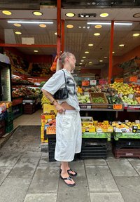 Fehér, laza szabású rövidnadrág és egy szürke, over size t-shirt fekete grafikával. Fekete kézitáska, strandszandál. Színes gyümölcsös stand a háttérben.