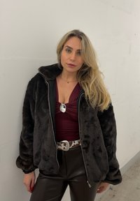 Femme blonde portant une veste en fausse fourrure noire, un haut bordeaux avec une boucle argentée, une ceinture à motif de vache, et un pantalon en cuir noir, se tenant contre un mur blanc.