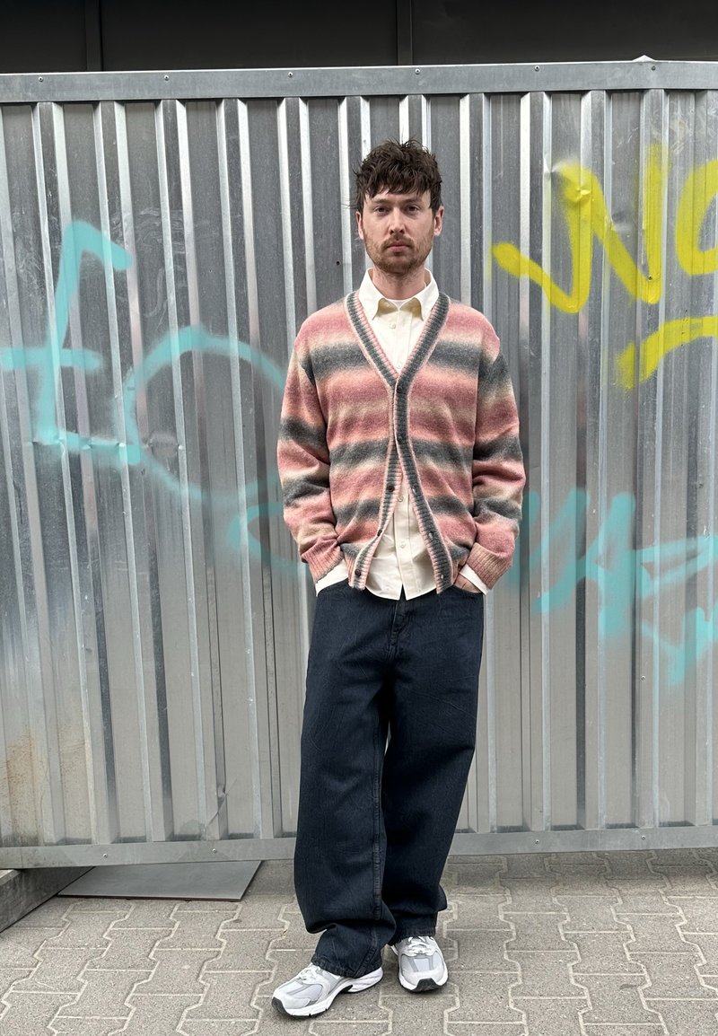 Homme aux cheveux foncés se tient contre un mur métallique couvert de graffitis, portant un cardigan rayé rose et gris, une chemise blanche, un pantalon noir large et des baskets blanches.