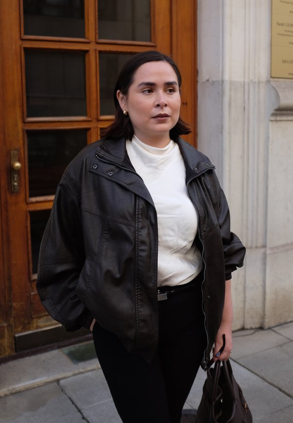 Veste en cuir noir avec fermeture éclair, portée par-dessus un col roulé blanc. Un pantalon noir et un sac à main marron complètent la tenue.