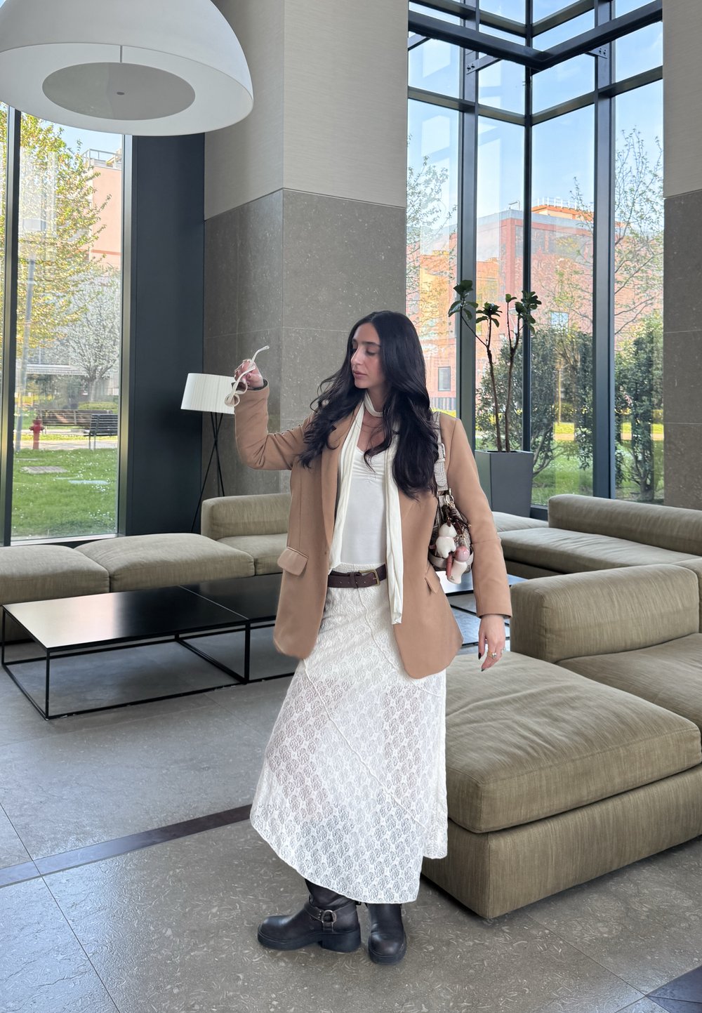 Καφέ υπερμεγέθη σακάκι, λευκό φόρεμα με δαντέλα, μαύρες μπότες, κρατώντας γυαλιά ηλίου. Σύγχρον�ος εσωτερικός χώρος με μεγάλες κουρτίνες, ορατή βλάστηση έξω.
