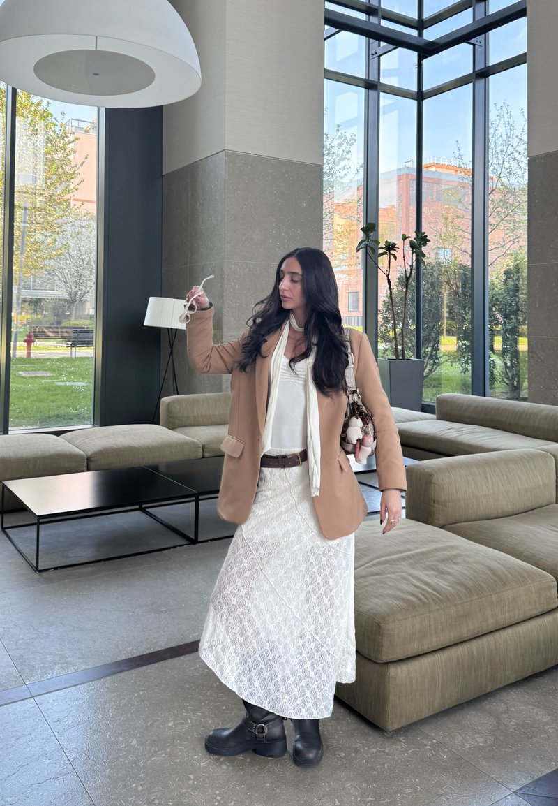 Blazer ligero marrón de gran tamaño, vestido de encaje blanco, botas negras, sosteniendo gafas de sol. Interior moderno con grandes ventanales, con vegetación visible afuera.