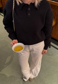 Persona vistiendo una camisa negra de manga larga y pantalones blancos, sosteniendo una taza de té claro en un ambiente interior informal.