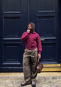 Man wearing a burgundy jumper, brown hood, wide beige trousers, and carrying a brown handbag stands against a dark blue paneled door.