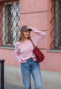 Roza pletena jopica, modri hlače z visokim pasom, rjava kapa; držanje rdeče torbe. Ozadje obsega roza steno z dekorativnimi kovinskimi žicami.