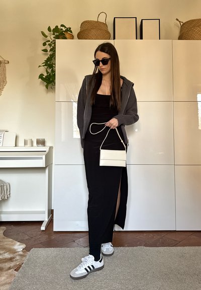 Mujer con vestido negro con abertura lateral, suéter gris con cremallera, zapatillas blancas, gafas de sol oscuras, sosteniendo un pequeño bolso blanco en una habitación moderna.