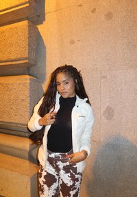 Λευκό φουσκωτό μπουφάν, μαύρο πουλόβερ, παντελόνι καφέ με άσπρο σχέδιο, ασημένια δαχτυλίδια, στέκεται μπροστά σε μια υφή γκρι τοίχο τη νύχτα.