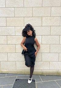 Svart ärmlös klänning med en strukturerad finish, knälång, i kombination med svarta strumpbyxor och vita ballerina skor. Enkel svart axelremsväska.