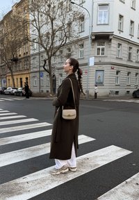 Manteau long olive, pantalon blanc et baskets beige, avec un petit sac bandoulière. Debout sur un passage pour piétons, fond urbain avec des arbres.