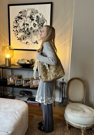 Braune, geflochtene Tragetasche mit dekorativen Spitzenakzenten, die über der Schulter getragen wird. Der Hintergrund zeigt ein rundes schwarz-weißes Kunstwerk und eine leuchtende Lampe.