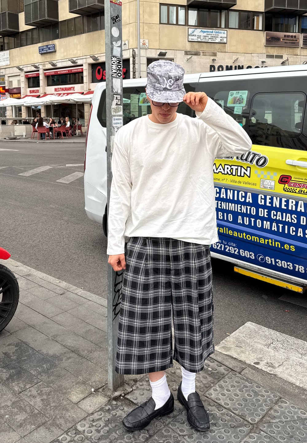 Valge pikad varrukad särk, must-valge ruuduline culottes, hall kamuflaažiga kübara, mustad loaferid ja valged sokid, seistes tänava post kõrval.