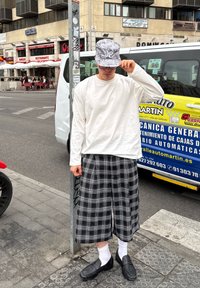 Valge pikad varrukad särk, must-valge ruuduline culottes, hall kamuflaažiga kübara, mustad loaferid ja valged sokid, seistes tänava post kõrval.