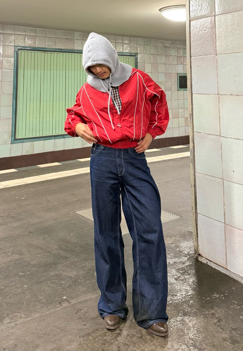 Veste en nylon rouge avec des rayures blanches, une capuche grise et une chemise à carreaux noire en dessous. Jean en denim foncé ample et chaussures marron.