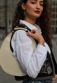 Hobo torba iz kremnega usnja s zaobljenimi oblikami in črnimi akcenti. Model nosi belo srajco in vezeno črno brezrokavnik.