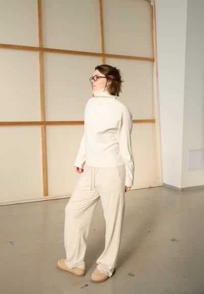 Persona con gafas y atuendo color crema con cuello alto, de pie en el interior sobre un suelo gris, frente a un fondo de marco de madera clara.