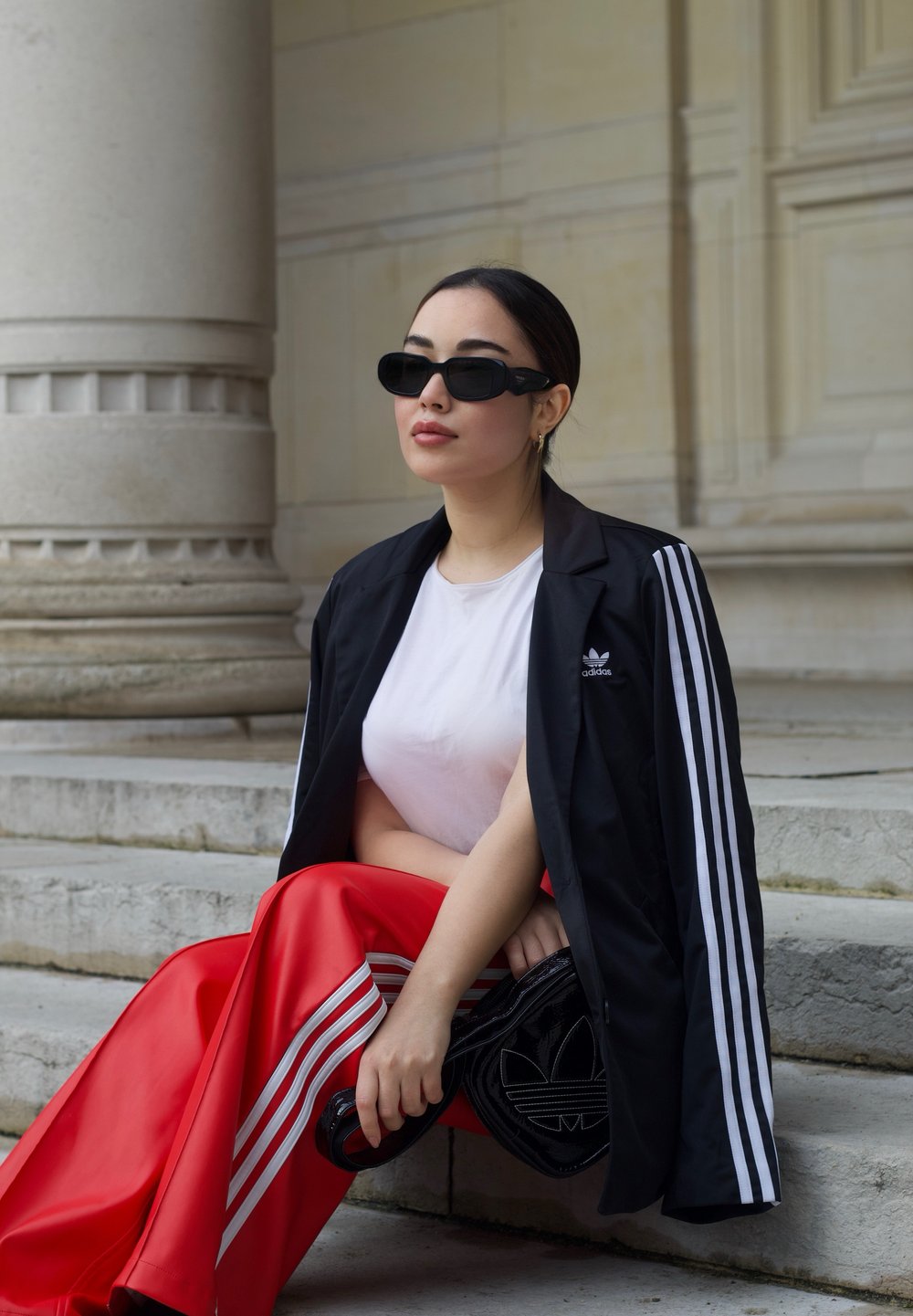 Veste noire Adidas avec des rayures blanches, t-shirt blanc, pantalon rouge brillant et lunettes de soleil noires ; tenant un sac noir avec le logo Adidas.