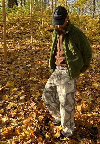 Chaqueta de forro polar verde sobre una camisa marrón, pantalones de camuflaje y gorra negra. Rodeado de hojas de otoño en una zona boscoso.