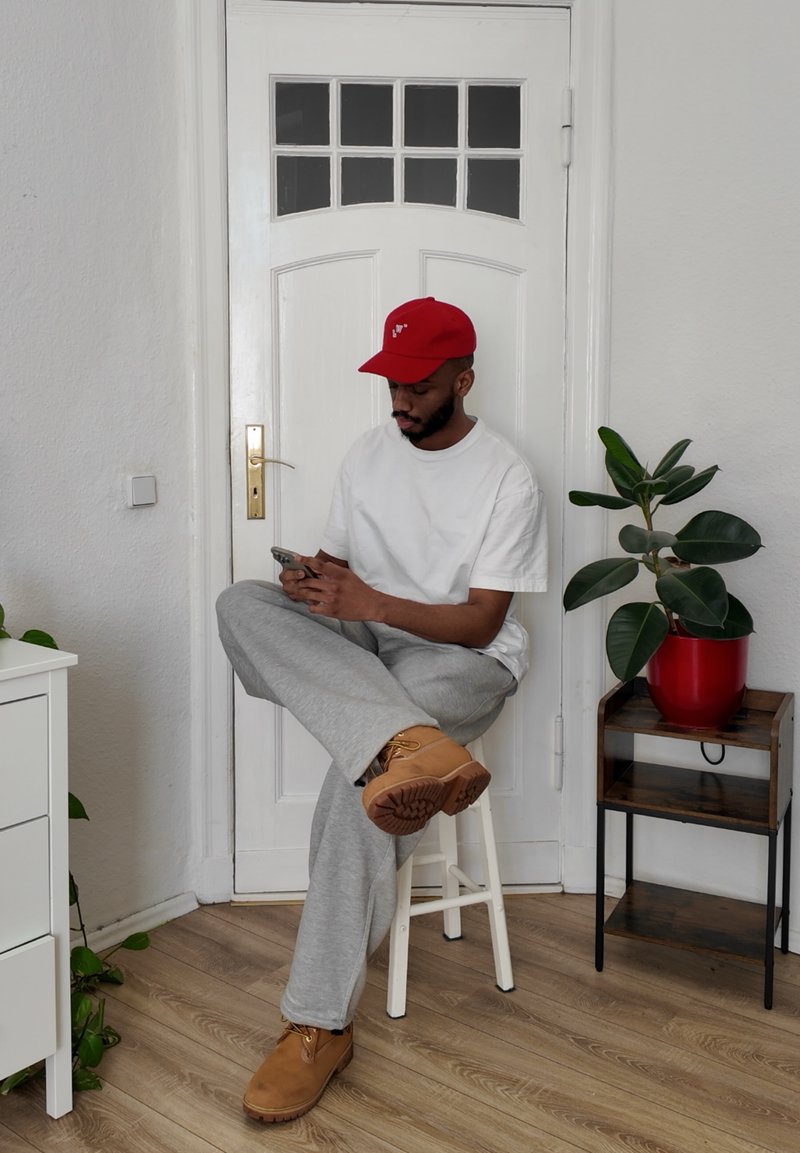 En mand med en rød kasket sidder på en hvid skammel, iført en hvid skjorte og grå sweatpants. Tan støvler er synlige. En potteplante er i nærheden.
