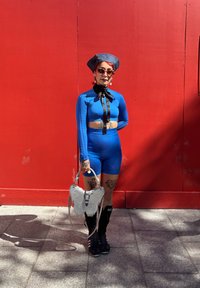 Persona con capelli rossi indossa un crop top a maniche lunghe blu e pantaloni corti, stivali neri, un cappello blu, occhiali da sole e tiene in mano una borsa a forma di farfalla bianca.