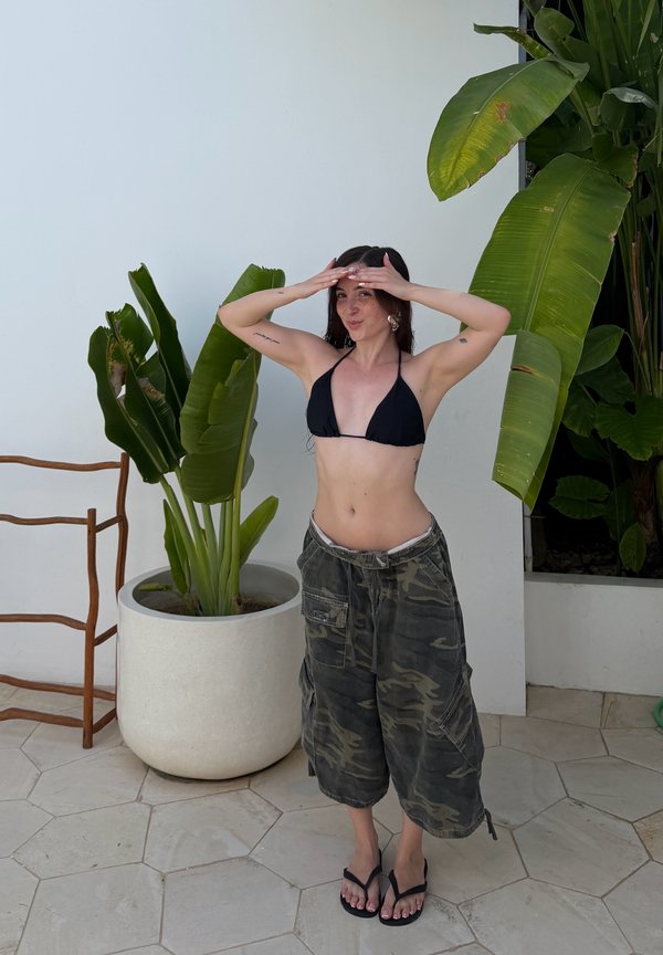 Kobieta w czarnym topie bikini, spodniach moro i klapkach pozująca w pomieszczeniu obok dużej doniczki z tropikalną rośliną i drewnianego krzesła.